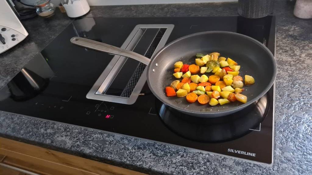 Nahaufnahme von oben auf eine graue Küchenarbeitsplatte inklusive einer Pfanne mit buntem Gemüse auf einem schwarzen Silverline Induktionskochfeld im Test, auf dem sich ein mittiger Dunstabzug befindet.