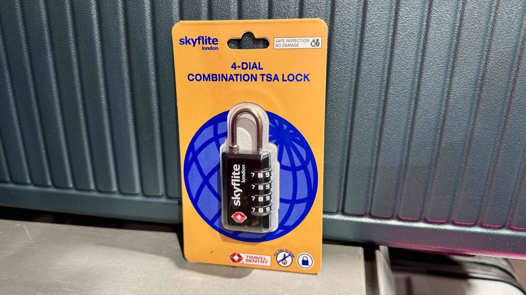 Ein Kofferschloss im Test lehnt innerhalb seiner Verpackung an einem grauen Koffer.