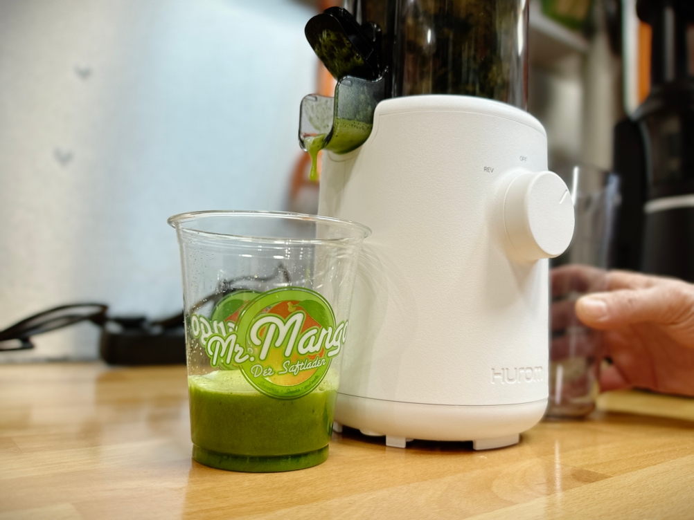 Ein Glas mit frisch gepresstem Saft steht neben dem Hurom H310A, während eine Hand nach einem Trester-Behälter greift.