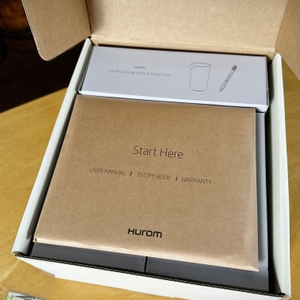 Der geöffnete Karton des Hurom H310A Slow Juicers zeigt die enthaltenen Einzelteile. Die Verpackung ist sauber und geordnet gestaltet.