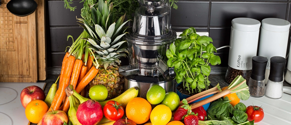 Ein Slow-Juicer steht in einer Küche, umgeben von zahlreichem Obst und Gemüse
