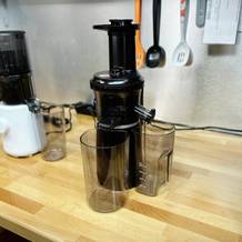 Der Panasonic MJ-L501KXE Slow Juicer steht zusammen mit seinem Zubehör auf einer Arbeitsplatte. Im Hintergrund befindet sich ein weiterer Slow Juicer in Weiß.