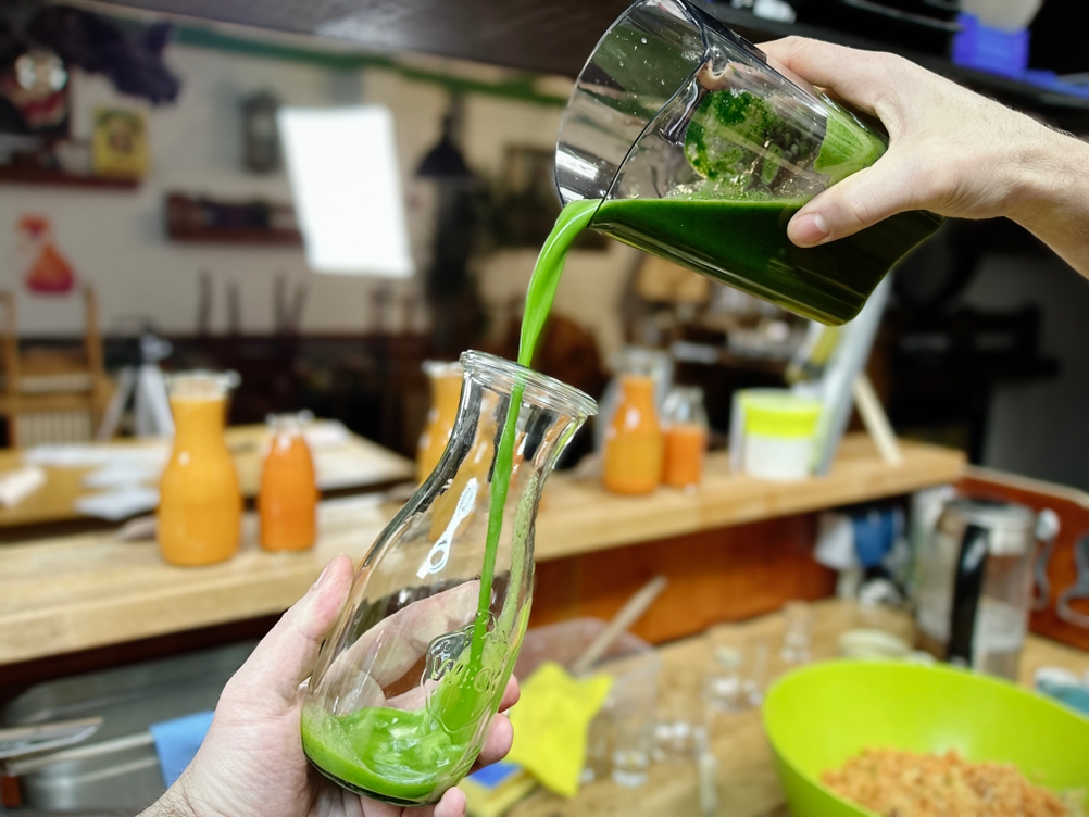 Frischer grüner Saft wird aus einem Behälter in eine Glasflasche gegossen. Im Hintergrund stehen mehrere gefüllte Saftflaschen.