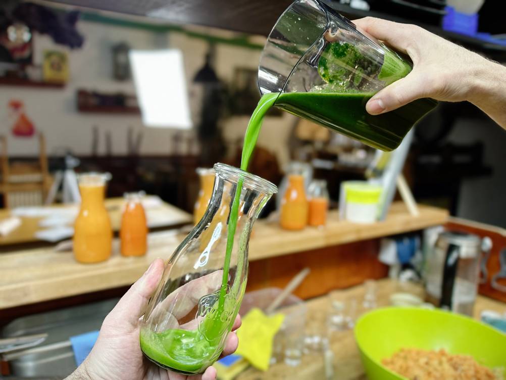 Frischer grüner Saft wird aus einem Behälter in eine Glasflasche gegossen. Im Hintergrund stehen mehrere gefüllte Saftflaschen.