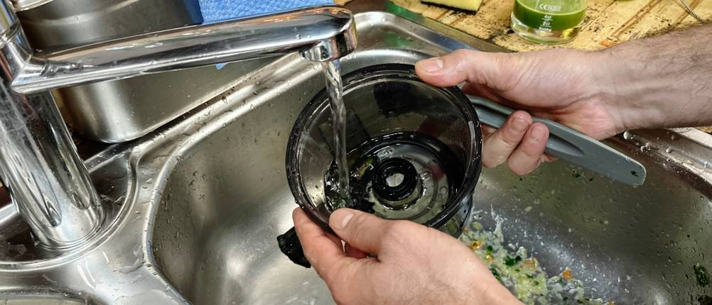 Ein Mann spült ein Bauteil des Panasonic MJ-L501KXE unter fließendem Wasser in einer Küchenspüle. Im Hintergrund liegen Reinigungstücher und Küchenutensilien.
