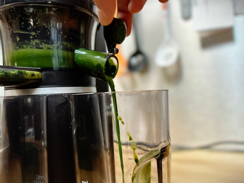 Grüner Saft fließt aus dem Auslauf des Panasonic MJ-L501KXE in einen Auffangbehälter. Im Hintergrund sind Küchenutensilien zu sehen.