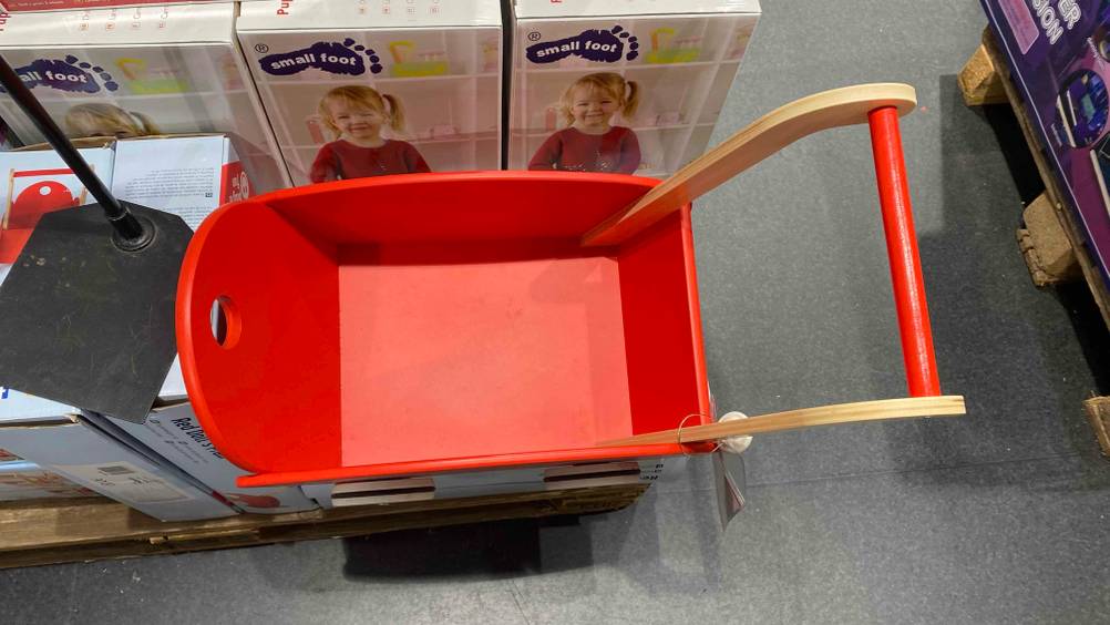 Ein getesteter Puppenwagen aus Holz von oben auf einer Holzpalette vor mehreren verpackten Produkten in einer Ausstellungsfläche.