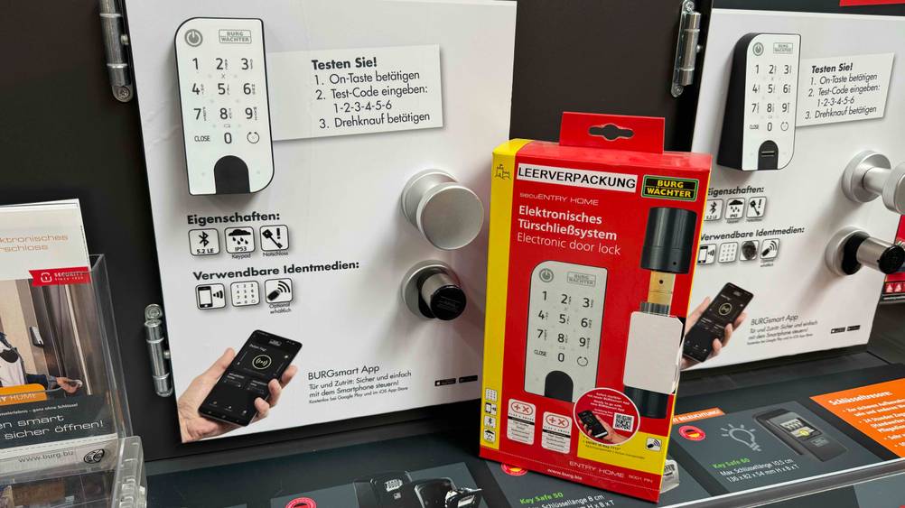 Ein getestetes smartes Türschloss montiert an einer Infotafel, daneben ein originalverpacktes Gerät.