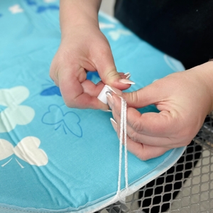 Eine Person fixiert den Kordelzug des smartgentle-Bügeltischbezugs mithilfe eines weißen Stoppers. Der Bezug liegt glatt auf dem Bügelbrett.