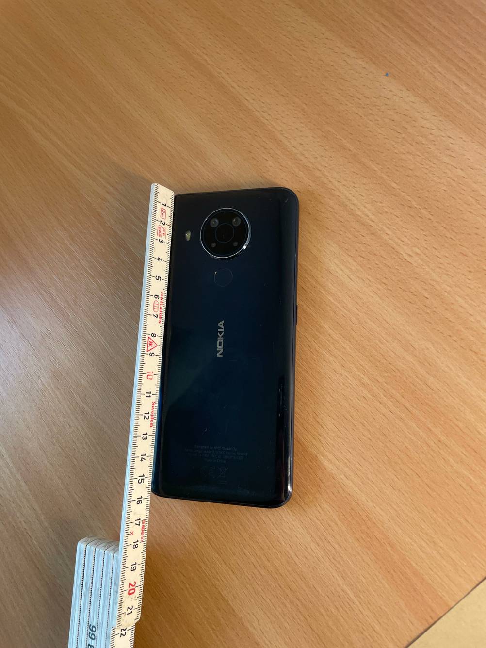 Ein getestetes Smartphone bis 300 Euro liegt mit der Rückseite nach oben auf einer Holzfläche an der Seite befindet sich ein Meterstab.