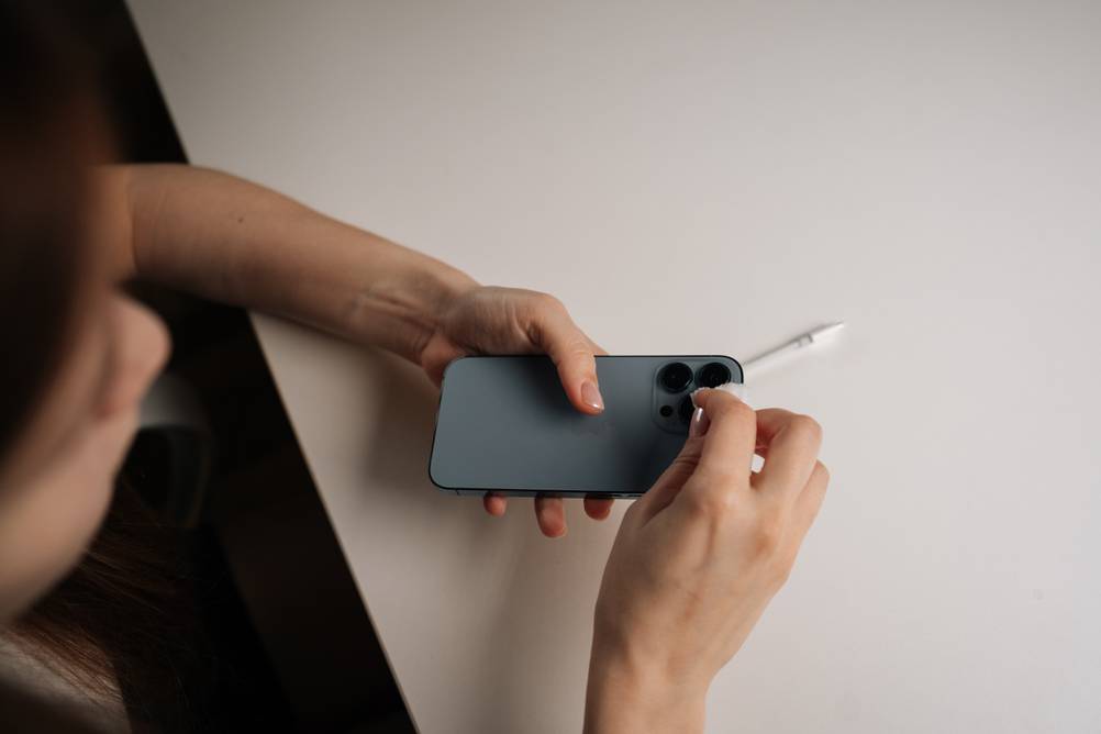 Eine Frau reinigt behutsam den Kamerabereich eines Smartphones. Sie blickt dabei von oben herab auf das Smartphone, dass sie in den Händen hält. Dafür benutzt sie ein kleines Tuch. Im Hintergrund ist eine Tischplatte zu sehen, auf der ein Ladekabel liegt.