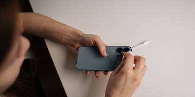 Eine Frau reinigt behutsam den Kamerabereich eines Smartphones. Sie blickt dabei von oben herab auf das Smartphone, dass sie in den Händen hält. Dafür benutzt sie ein kleines Tuch. Im Hintergrund ist eine Tischplatte zu sehen, auf der ein Ladekabel liegt.