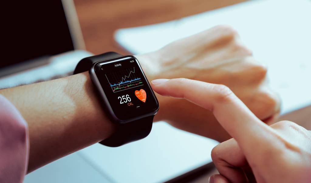Eine Frau trägt an ihrem Handgelenk eine Smartwatch mit verschiedenen Werten, die darauf angezeigt werden. Sie tippt mit dem Finger auf die Uhr; im Hintergrund ist ein Laptop zu sehen.