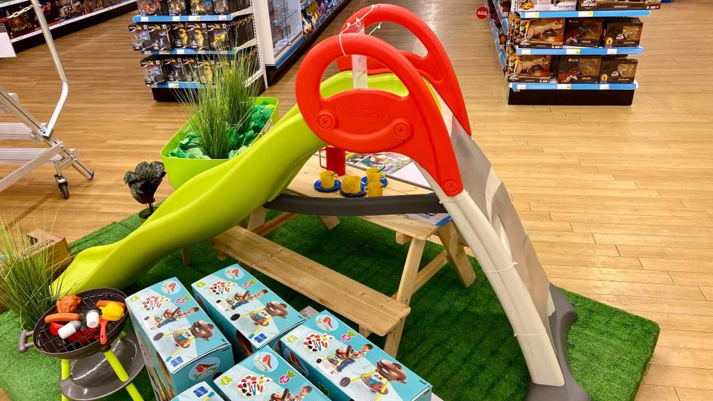 Kinderrutsche im Test: Ein Modell aus Kunststoff mit hellgrüner Rutsche, rotem Geländer und beigefarbener Treppe steht auf einem Kunstrasen neben Spielzeugpackungen in einem Geschäft.
