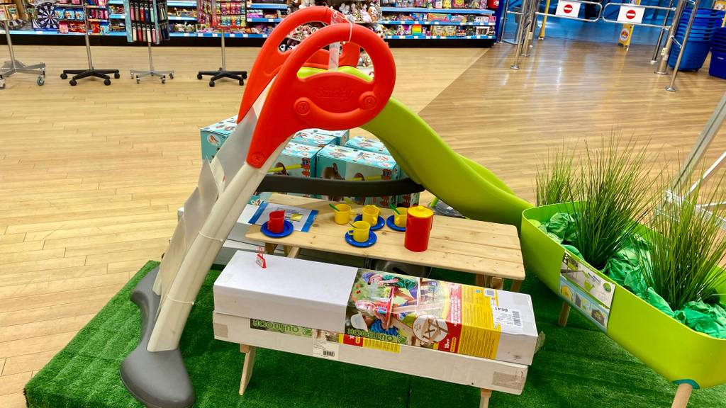 Kinderrutschen-Test: eine Anfertigung aus Kunststoff mit hellgrüner Rutsche, rotem Geländer und beigefarbener Treppe befindet sich in einem Spielwaren-Verkaufsbereich hinter seinem Verpackungskarton auf einem Kunstrasen.
