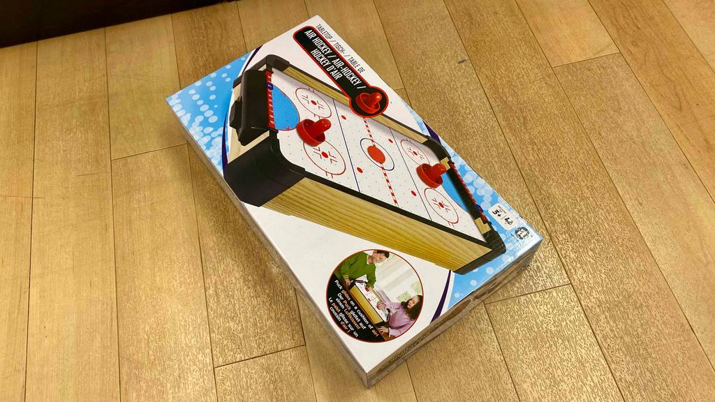 Originalverpackung eines getesteten Air Hockey liegt schräg auf einem Holzboden.