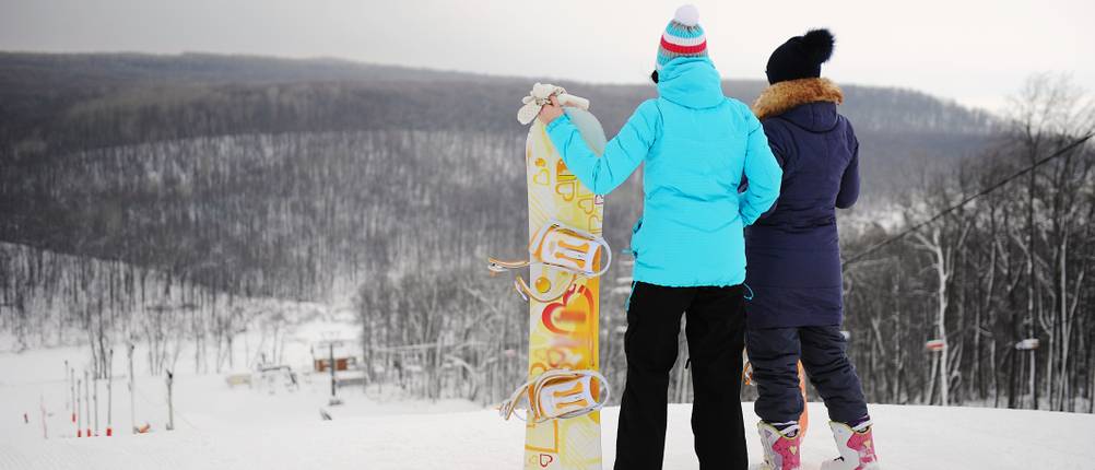 Snowboardtaschen-Test: zwei Personen, die mit ihren Snowboards auf Fuße einer Abfahrtspiste stehen