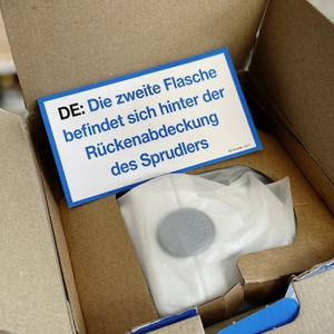 Geöffnete Verpackung des SodaStream Cool mit einem Hinweis, dass die zweite Flasche hinter der Rückenabdeckung des Sprudlers versteckt ist.