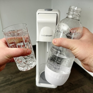 Eine Hand hält ein Glas mit sprudelndem Wasser, die andere eine Kunststoffflasche. Im Hintergrund steht der SodaStream Cool.