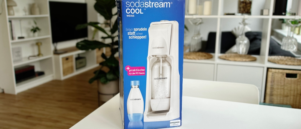 Die Verpackung des SodaStream Cool steht auf einem weißen Tisch mit moderner Einrichtung im Hintergrund.
