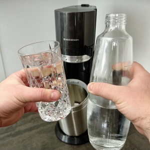 Eine Hand hält ein Glas mit sprudelndem Wasser, die andere eine Glasflasche. Im Hintergrund steht der SodaStream Duo.