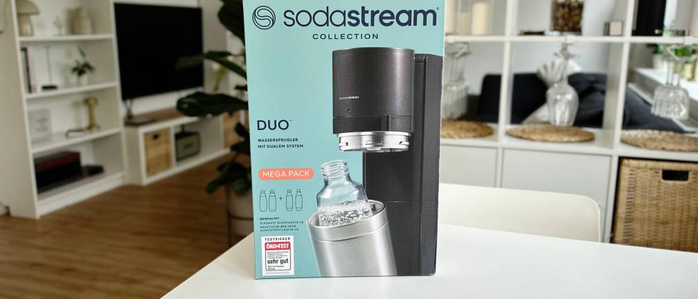 Der SodaStream Duo in seiner Verpackung auf einem weißen Tisch mit einer modernen Wohnungseinrichtung im Hintergrund.