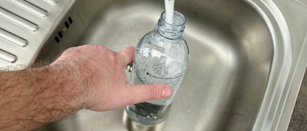 Eine Hand spült eine Glasflasche unter fließendem Wasser in einem Edelstahlspülbecken aus. Die Flasche gehört zu einem Wassersprudler.
