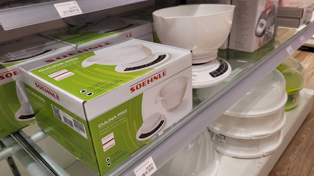 Eine getestete analoge Küchenwaage von Soehnle mit schwarzer Gewichtsskala und weißer Kunststoffschale steht auf einer Glasfläche neben seiner Originalverpackung.