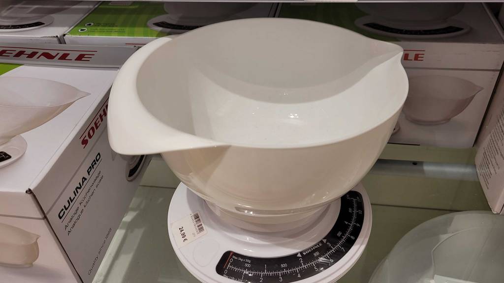 Ansicht von schräg oben auf eine weiße, getestete analoge Küchenwaage der Marke Soehnle inklusive Kunststoffschale sowie schwarzer Gewichtsskala auf einer Glasfläche neben Originalverpackungen derselben.