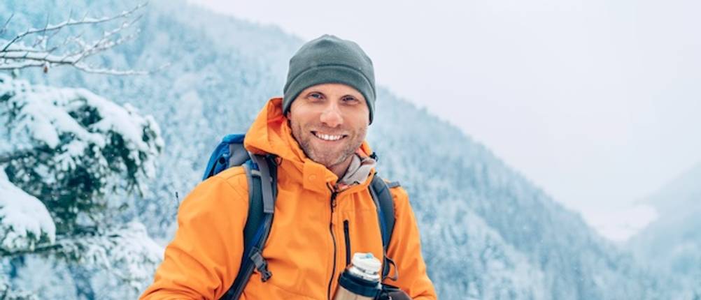 Getestete orangefarbene Softshelljacke für Herren, beim Wandern von einem Herrn mit Mütze und Rucksack getragen.