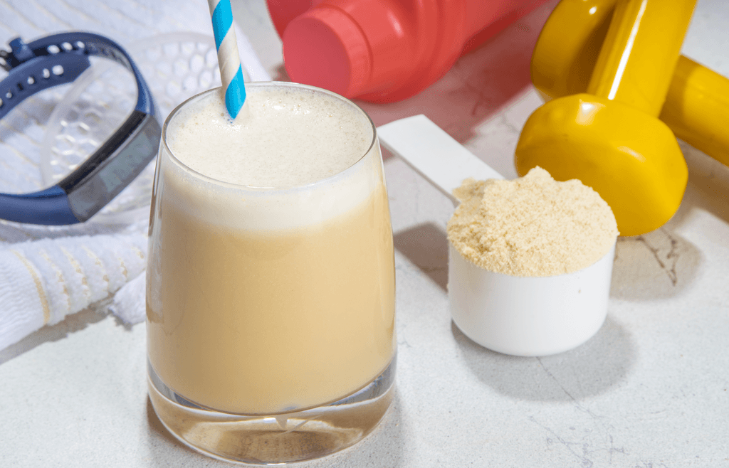 Soja-Protein-Test: Auf einem Tisch steht ein Soja-Protein-Shake in einem Glas mit Strohhalm. Daneben befindet sich ein Löffel mit Soja-Protein und Sportzubehör. 