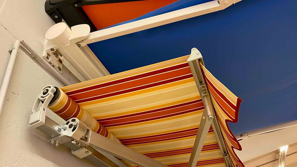 Nahaufnahme von ausgefahrenen Markisen in Orange, in Blau und in Gelborange an einer Ausstellungswand im Verkaufsbereich.