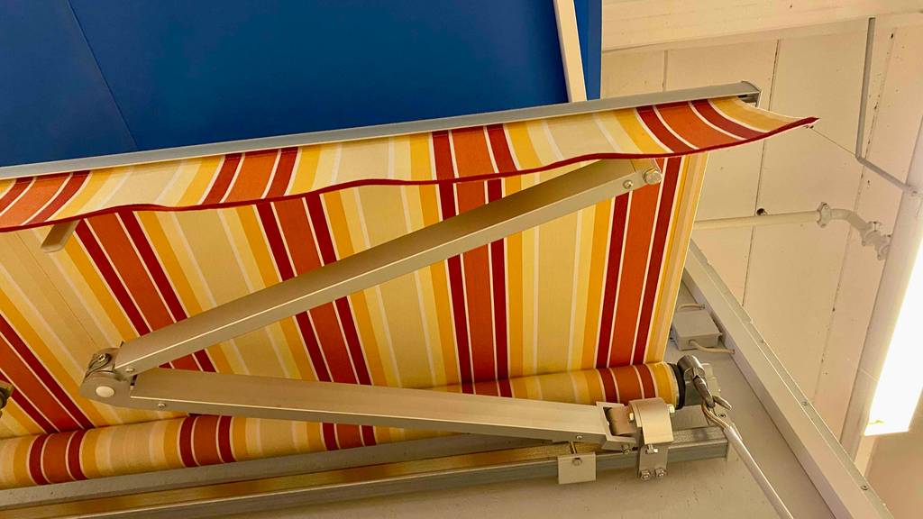Markise im Test: Detailansicht auf eine orangegelbe ausgezogene Markise, mit Fokus auf den Ein- und Ausfahrtmechanismus, ist an einer Ausstellungswand im Verkaufsbereich montiert.