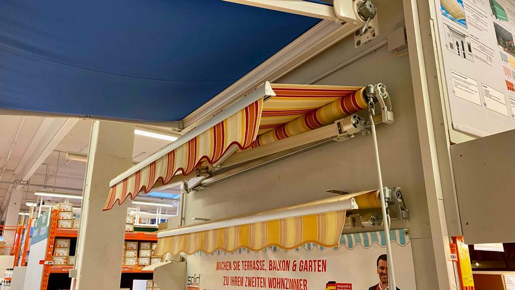 Markisen-Test: drei Terrassen-Modelle, in Blau, Orangegelb und Beige-Gelb gestreift befinden sich an einer Ausstellungswand montiert im Geschäft.