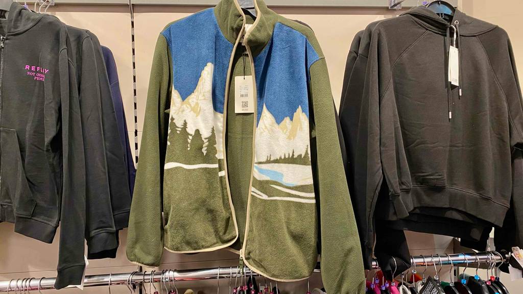 Herren-Fleecejacken im Test: Ein olivgrün-blau-weißes Modell mit Bergmotiv auf der Vorderseite hängt mit geöffnetem Reißverschluss zwischen grauen Baumwoll-Jacken an einer Wand im Verkauf.
