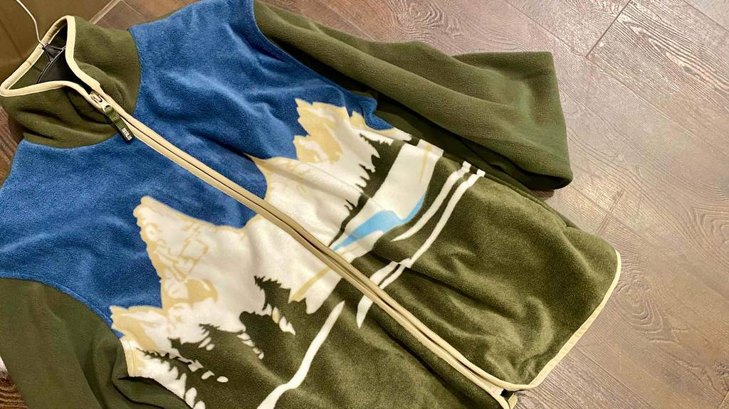 Fleecejacken für Herren im Test: Ein olivgrün-blau-weißes Modell mit Bergmotiv auf der Vorderseite inklusive Reißverschluss liegt auf einer Holzfläche.