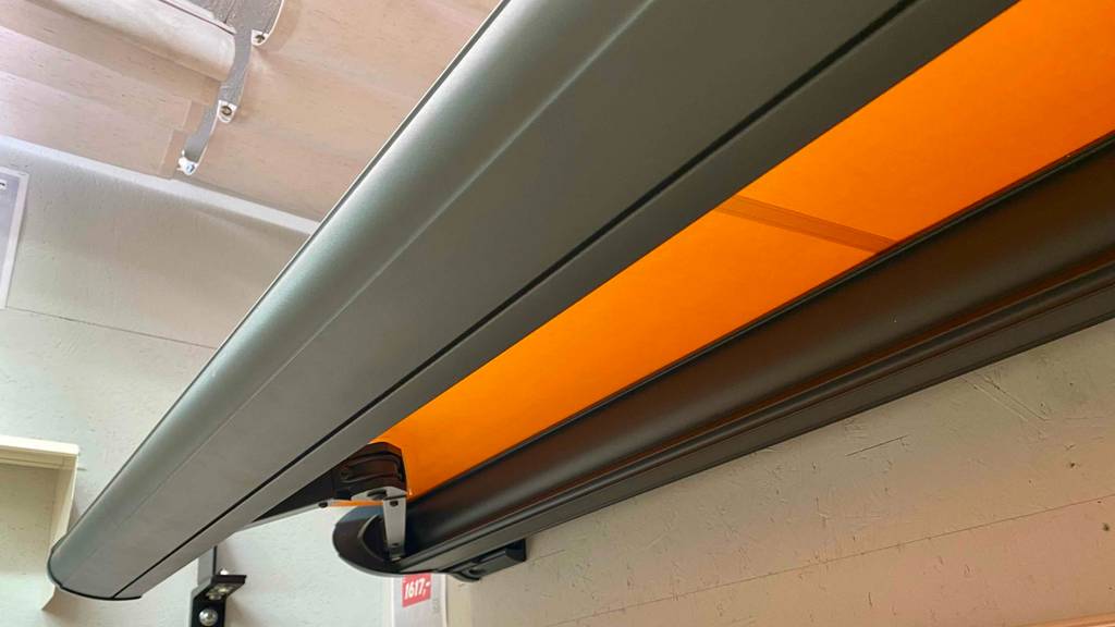 Kassettenmarkisen im Test: Detailansicht von der Seite auf den Ausfahrtmechanismus eines orangen Modells inklusive Preisschild an weißer Wand.