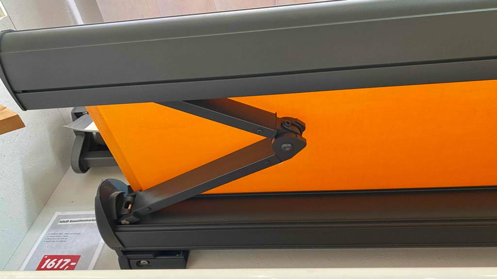 Kassettenmarkisen-Test: Detailansicht von dem Ausfahrtmechanismus eines orangen Modells neben seinem Preisschild.