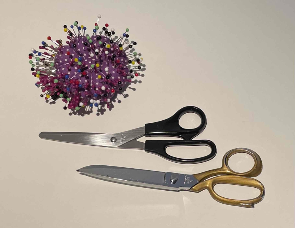 Schneiderscheren im Test: zwei Stoffscheren befinden sich neben einem Kissen mit Stecknadeln auf einer hellen Fläche.