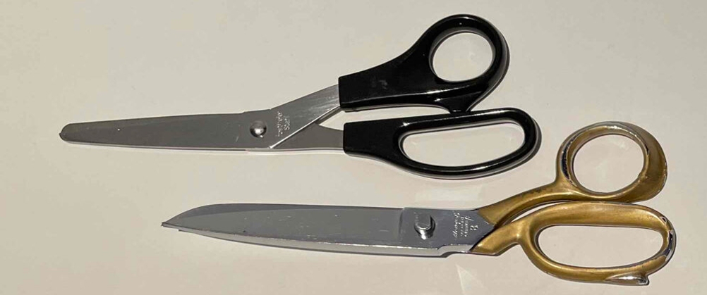 Zwei getestete Schneiderscheren, mit schwarzem und goldfarbenem Griff, liegen auf einer weißen Oberfläche.