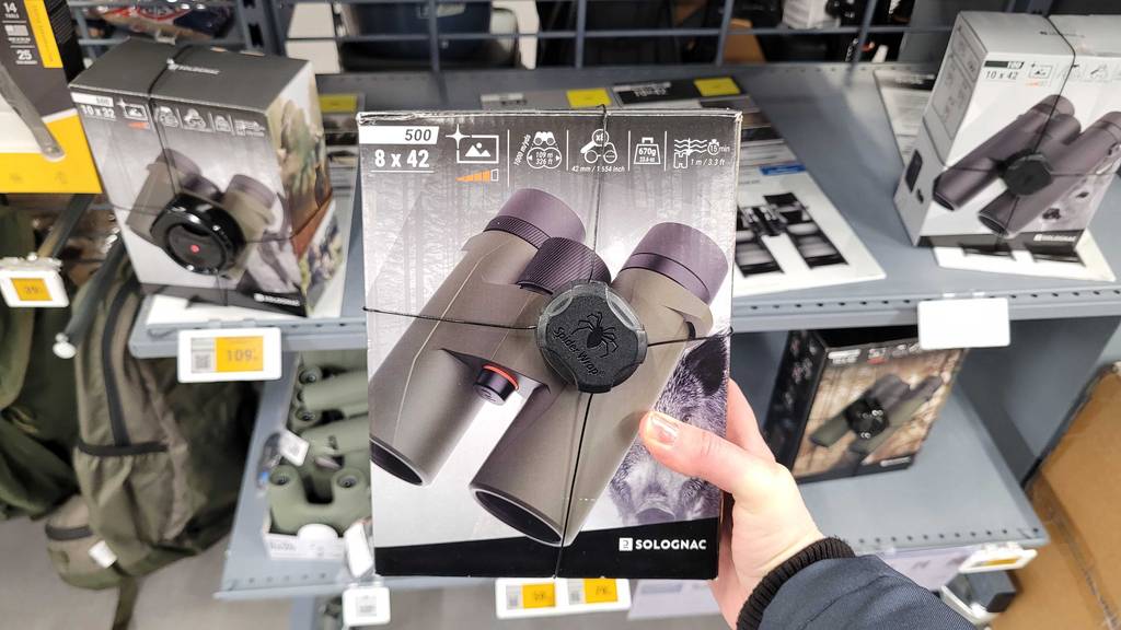 Eine Hand hält die gesicherte Verpackung eines getesteten 8-x-42-Fernglases vor einem Verkaufsregal.