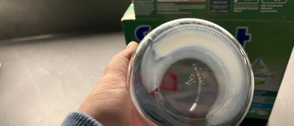 Eine Hand hält ein Glas mit eingetrockneter Milch fest. Im Hintergrund steht die Packung mit den Somat All-in-1 Pro Nature Spülmaschinentabs