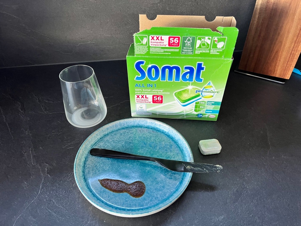 Der Karton mit den Somat All-in-1 Pro Nature Spülmaschinentabs liegt hinter einem Tab, einem Teller mit eingetrocknetem Ketchup, einem Glas mit eingetrockneter Milch und einem Messer mit Butter