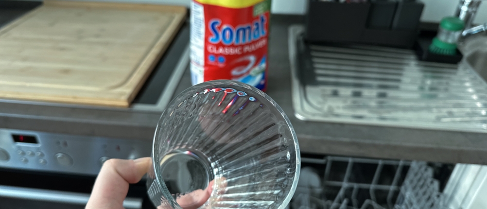 Ein sauberes Glas wird vor einer offenen Spülmaschine hochgehalten.