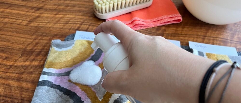 Eine Hand gibt etwas Sonax XTREME Polsterreiniger auf einen Rotweinfleck auf einem Kissenbezug.