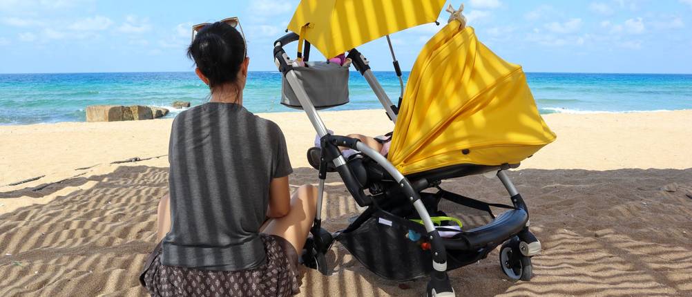 Sonnenschirm kinderwagen test: Frau sitzt mit gelbem Kinderwagen mit Sonnenschirm am Strand