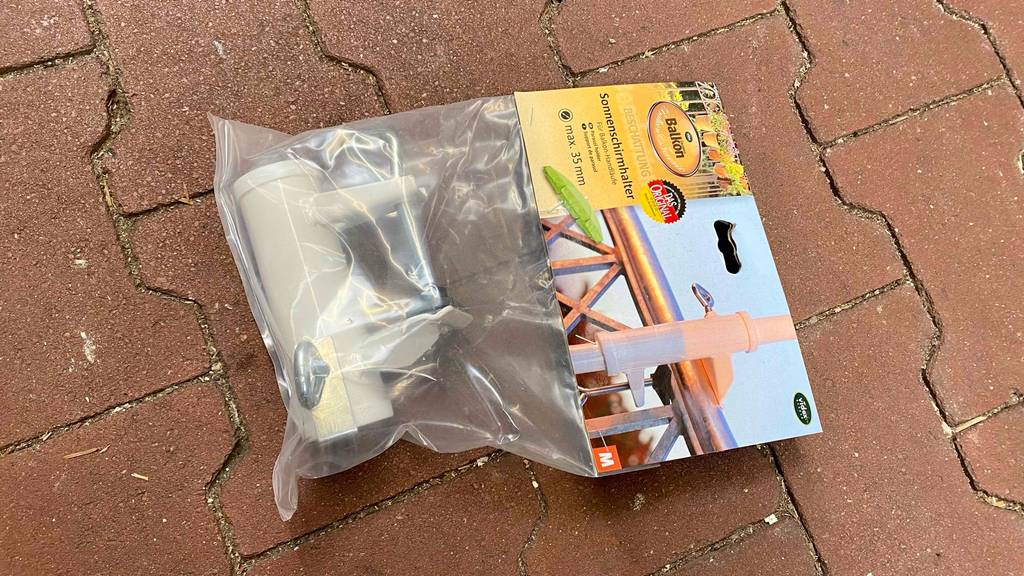 Ein getesteter Sonnenschirmhalter für Balkongeländer liegt verpackt auf dem Boden.