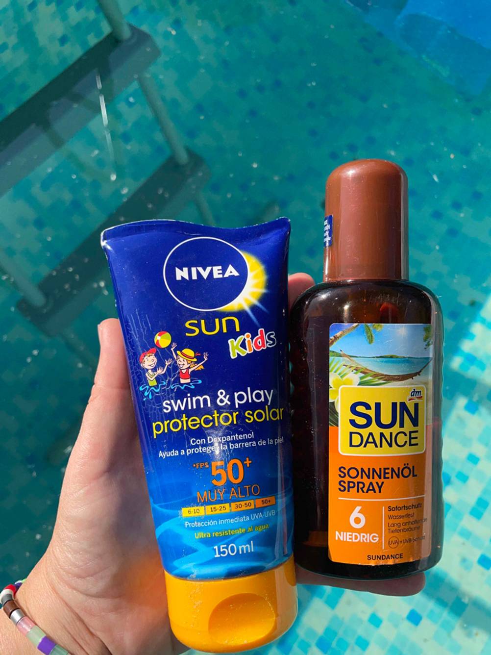 Sonnenschutzmittel im Test: Eine Hand hält zwei Sonnenschutzmittel vor einen Pool.