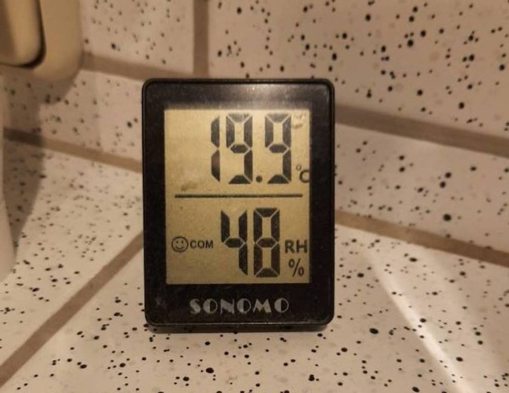 Hygrometer-Test: schwarzes Sonomo Modell mit eingeschaltetem LCD-Bildschirm steht auf weißen, schwarz gepunkteten Fliesen.