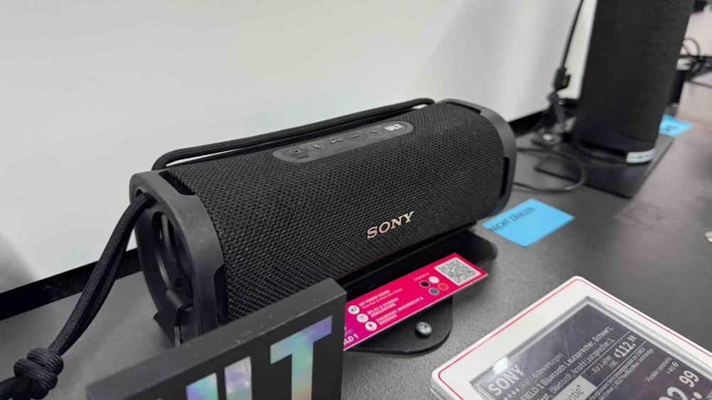 Ein Sony-Lautsprecher im Test auf einem Verkaufsregal neben anderen Geräten, davor ein Preisschild.